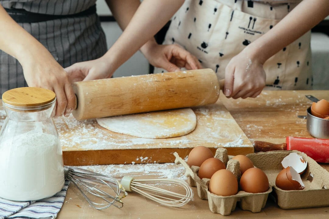 Hands rolling dough with a wooden rolling pin on a floured surface, surrounded by baking ingredients including eggs, flour, and a whisk, perfect for a baking blog post