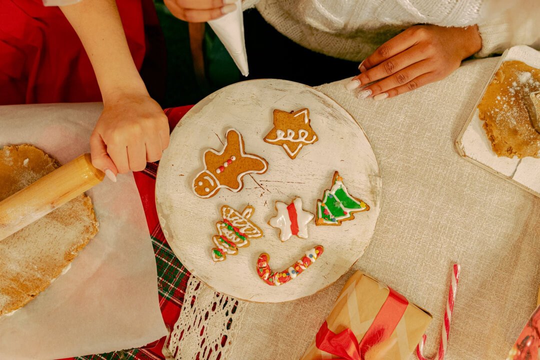 Hands decorating festive gingerbread cookies, including a gingerbread man and Christmas tree, on a wooden platter, with baking tools and holiday gifts in the background