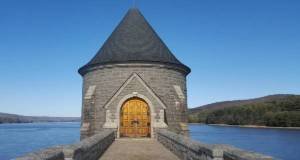 Stone tower with a conical roof and arched wooden doors stands on a dam overlooking a lake
